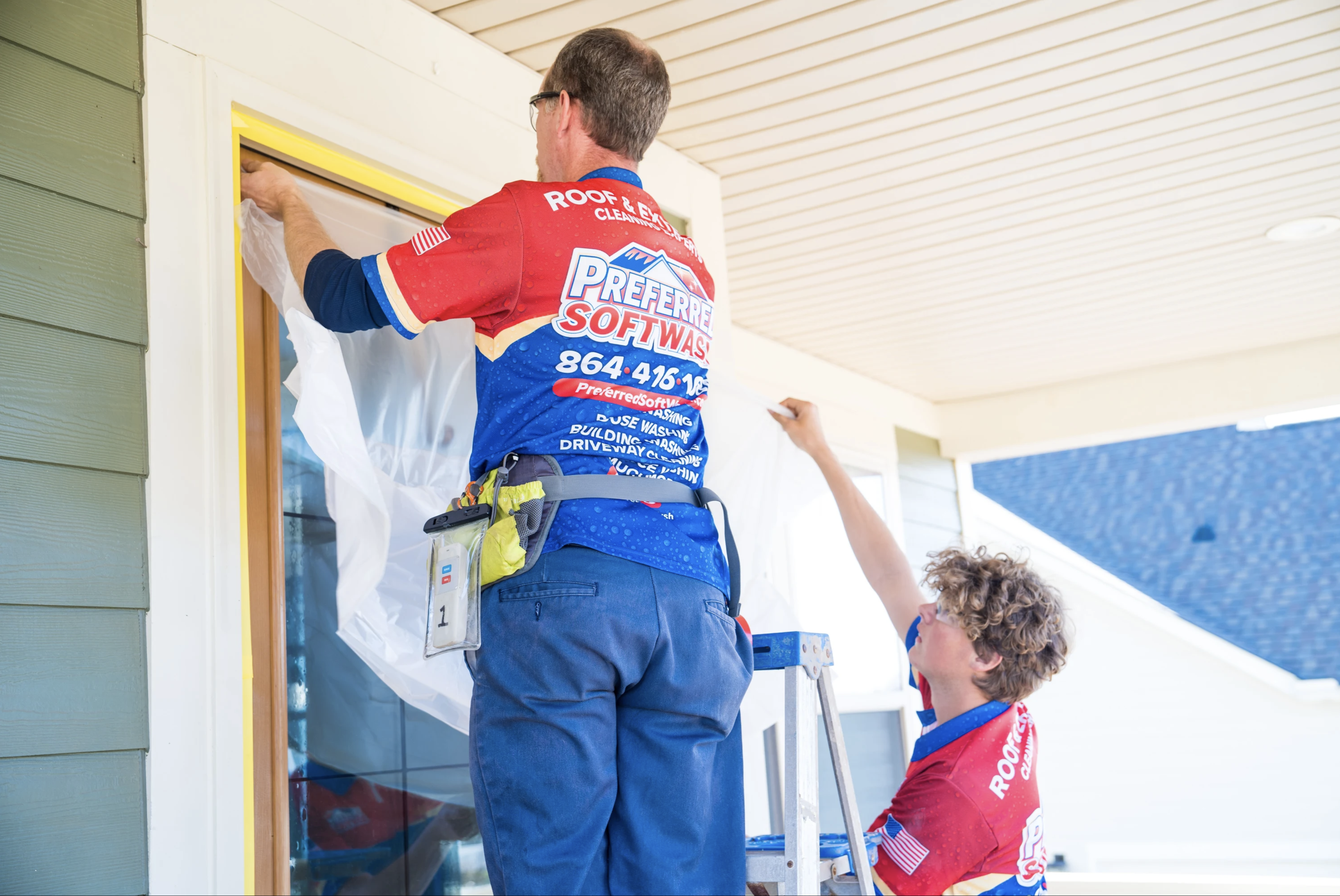 Two workers in red shirts from "Preferred Softwash" are preparing to clean the exterior of a house. One stands on a ladder, securing a plastic sheet over a window, while the other holds the sheet in place. The house has green siding and a wooden ceiling on the porch.